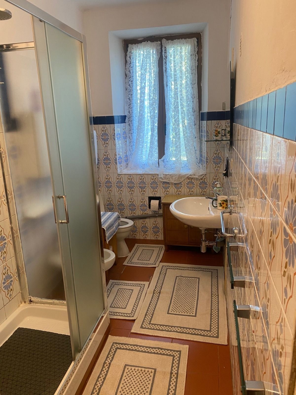 Bagno con doccia, piastrelle decorate e finestra con tende di pizzo